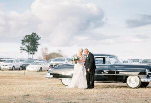 Bride standing beside her father and vintage car before an outdoor private estate wedding ceremony